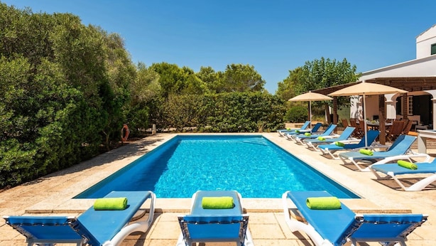 A rectangular swimming pool with blue water, surrounded by lounge chairs with blue cushions and green towels. - Villa Biniparrell by Villa Plus (Biniparrell)