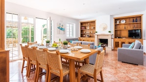 A spacious living area with a dining table set for six, surrounded by wicker chairs. A fireplace and a television are visible in the background. - Villa Biniparrell by Villa Plus (Biniparrell)
