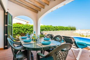 An outdoor dining area with a pool view, featuring a round table with chairs, a bottle of wine, and a glass. - Villa Blanc Arenal by Villa Plus (Es Mercadal)