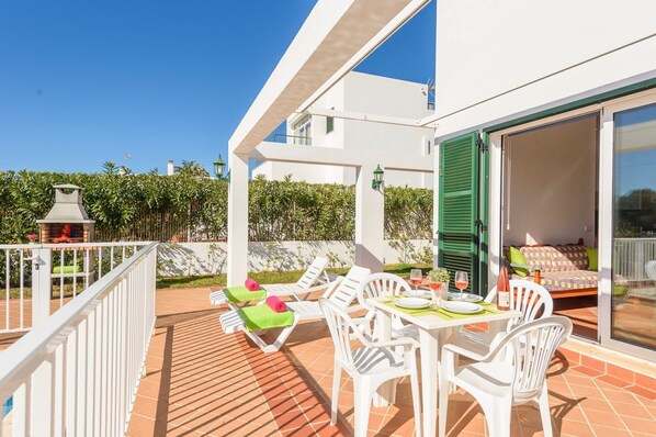 An outdoor patio with white furniture, a table set for dining, and a grill in the background.