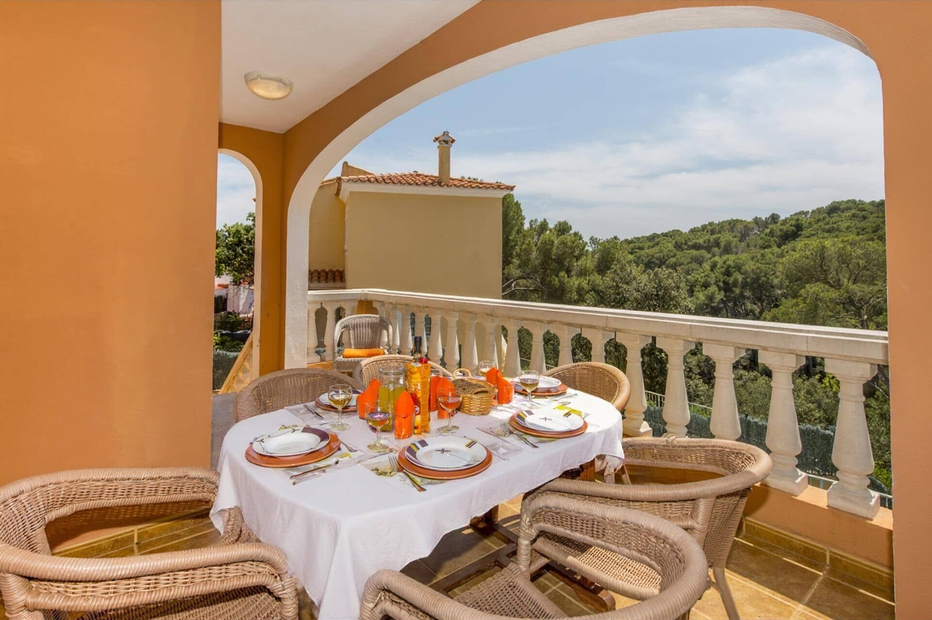 A dining table set for four with wicker chairs, a white tablecloth, and a view of a lush green landscape.