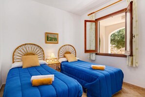 A room with two beds, each with a wooden headboard, a small bedside table with a lamp, and a window with curtains.