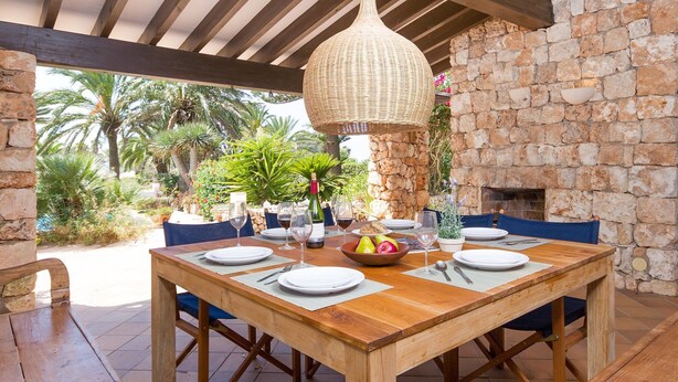 Un espace de restauration en plein air avec une table en bois prévue pour quatre personnes, entourée de murs en pierre et agrémentée d'un luminaire suspendu en osier.