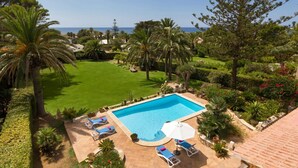 Une piscine dotée de chaises longues et d’un parasol, entourée d’un jardin bien entretenu et de palmiers.