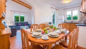 A dining area with a wooden table set for four, surrounded by wooden chairs. There is a sofa and a small table with a lamp in the background. - Villa Dolfi by Villa Plus (Los Delfines)