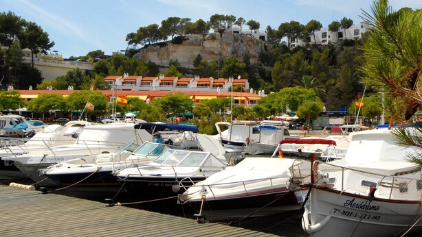 Un port de plaisance où sont amarrés plusieurs bateaux, un bâtiment avec des toits recouverts de tuiles rouges, et une falaise avec des arbres en arrière-plan.