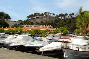 Un port de plaisance où sont amarrés plusieurs bateaux, un bâtiment avec des toits recouverts de tuiles rouges, et une falaise avec des arbres en arrière-plan.
