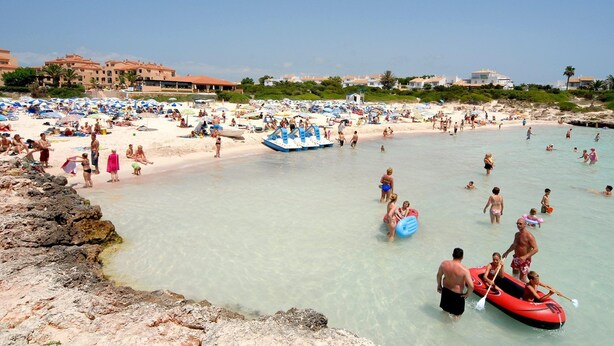 Ein belebter Strand mit badenden und sonnenbähenden Menschen, einem Rettungsturm und einer felsigen Küstenlinie.