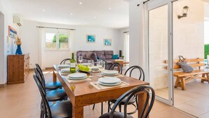 Un comedor con una mesa de madera dispuesta para cuatro personas, rodeada de sillas negras. En el fondo, hay un sofá y un banco, y un gran ventanal corredera que da a un área exterior.