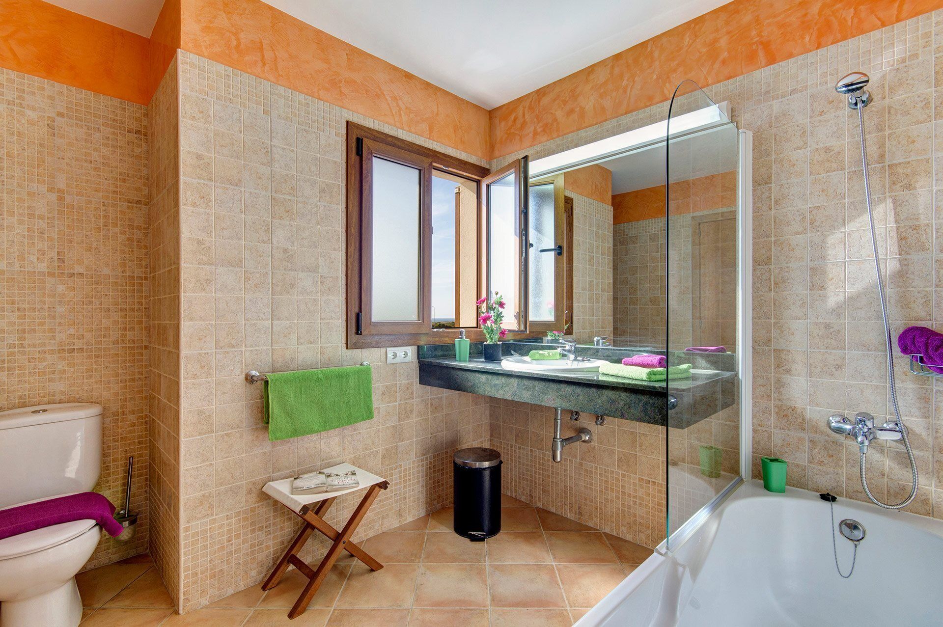 A bathroom with a toilet, a small table with a green towel, a glass shower enclosure, and a sink with a mirror above it.