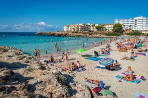 A beach with people sunbathing and swimming, a hotel in the background, and clear blue waters.