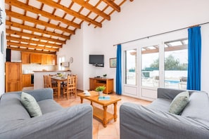 A spacious living room with a high ceiling, a dining area, and a view of a swimming pool through large glass doors. - Villa Ingrid Porter by Villa Plus (Cala en Porter)