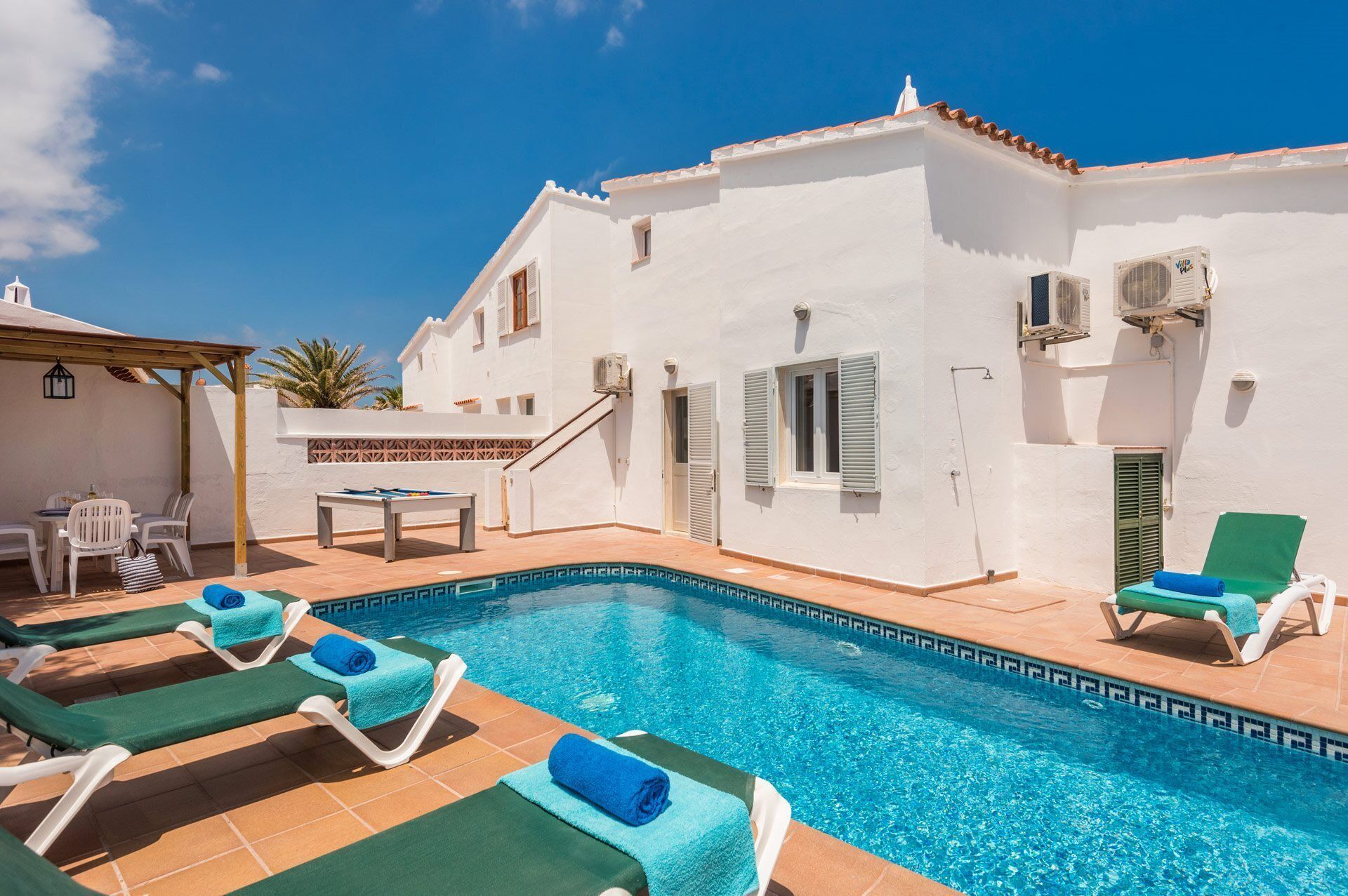 Un espace piscine avec des chaises longues et une table, un bâtiment blanc avec des volets, et un ciel bleu azur.