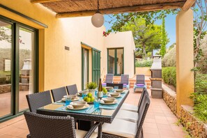 Un espace extérieur aménagé pour les repas, avec une table dressée pour quatre personnes, entourée de chaises en osier, et un barbecue à l’arrière-plan.