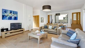 Un soggiorno moderno con una televisione a schermo piatto, un tavolino da caffè e un divano color beige. Sullo sfondo, una zona pranzo con tavolo e sedie.
