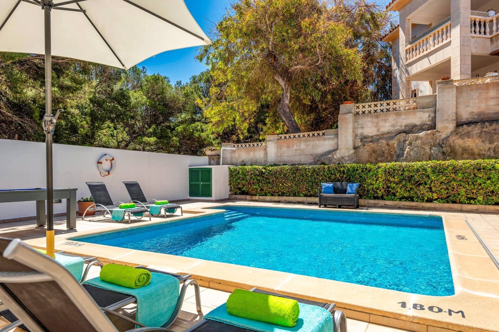 A swimming pool with lounge chairs and a table, surrounded by a low wall and greenery.