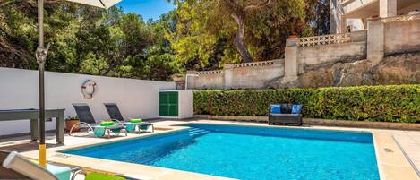 A swimming pool with lounge chairs and a table, surrounded by a low wall and greenery.