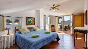 Ein Schlafzimmer mit Bett, Deckenventilator, Fenster, Tür und Blick ins Freie.