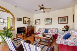 Un salon avec un téléviseur, un canapé, des fauteuils, un ventilateur de plafond et des tableaux encadrés accrochés au mur.