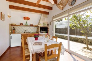Un coin repas avec une table dressée pour deux personnes, une cuisine dotée d’un four en briques et une vue sur un arbre à travers une porte vitrée.