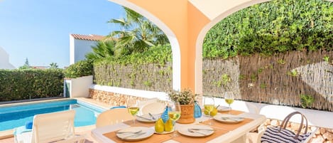 A dining area with a pool view, white furniture, and a tropical backdrop.