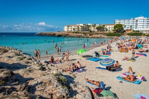 A beach with people sunbathing and swimming, a hotel in the background, and a rocky shoreline.