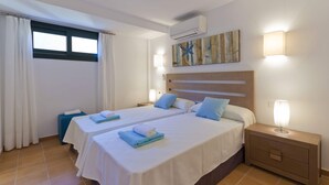 A hotel room with two beds, a window with curtains, an air conditioning unit, a bedside table with a lamp, and a wall-mounted artwork.
