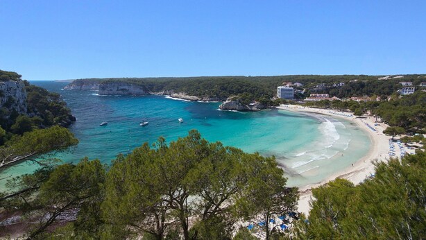 A coastal view with clear turquoise waters, a sandy beach, and a forested area.