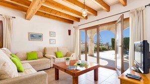 Una sala de estar con techo de madera, un sofá con cojines verdes, una mesa de centro y un televisor sobre un mueble de madera.