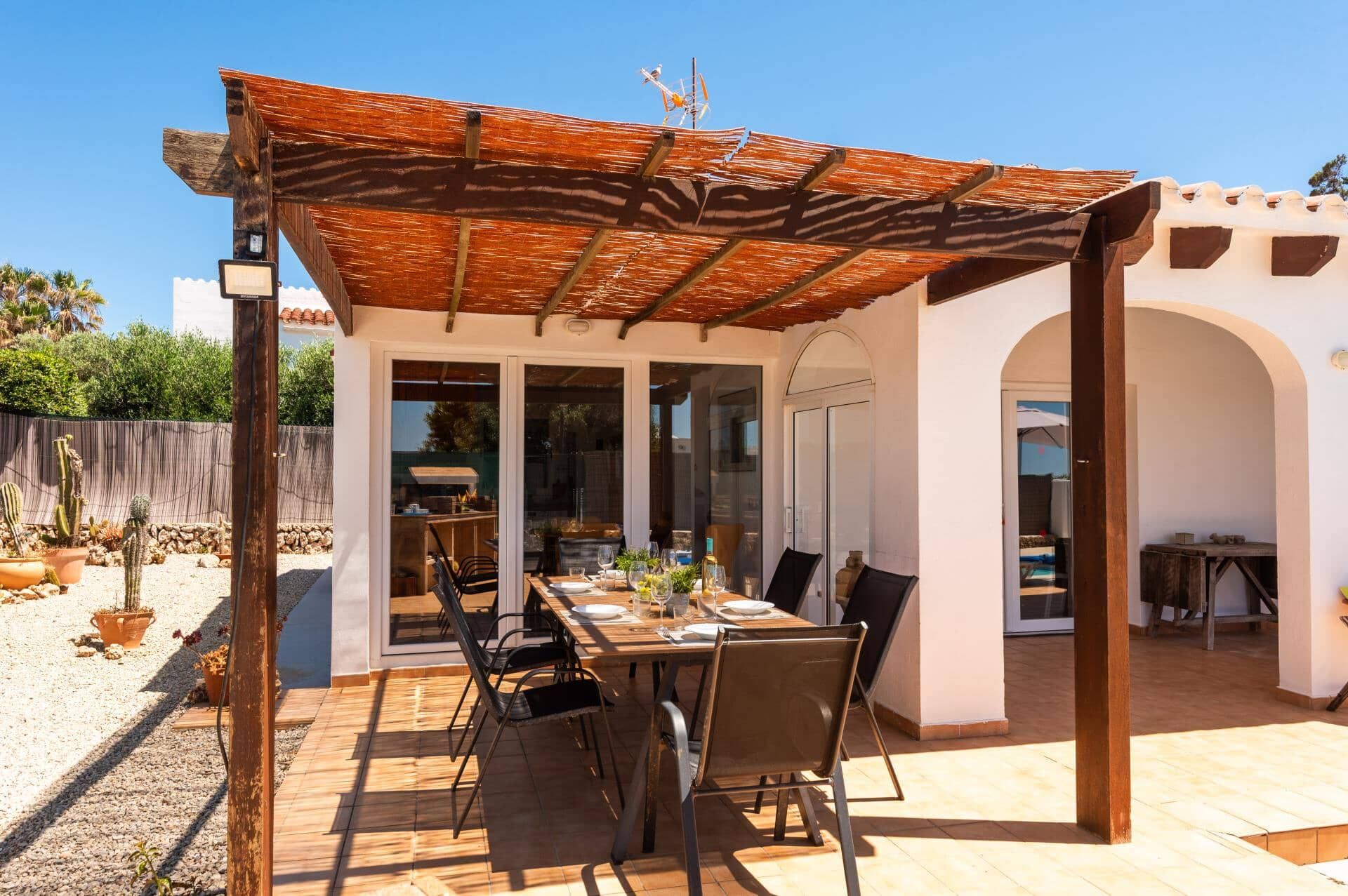 Une terrasse extérieure avec une table en bois dressée pour le repas, entourée de chaises, et un espace abrité avec un toit.