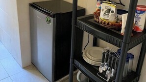 A compact kitchen with a microwave, a small refrigerator, and a metal shelf unit holding kitchenware and food items.