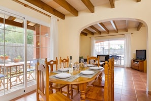 Un coin repas avec une table en bois dressée pour quatre personnes, un téléviseur en arrière-plan et une terrasse visible à travers des portes vitrées.