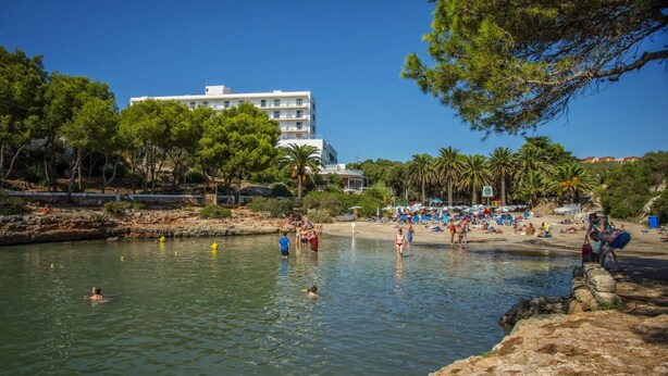 Una playa con agua cristalina, gente nadando y un edificio al fondo.