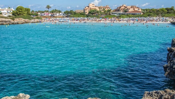 A coastal beach with clear turquoise water, a rocky shoreline, and buildings in the background. - Villa Noa by Villa Plus (Cap d'Artrutx)