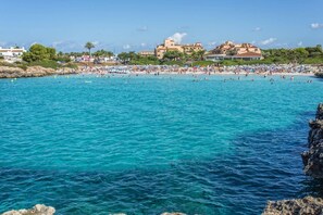 A coastal beach with clear turquoise water, a rocky shoreline, and buildings in the background. - Villa Noa by Villa Plus (Cap d'Artrutx)