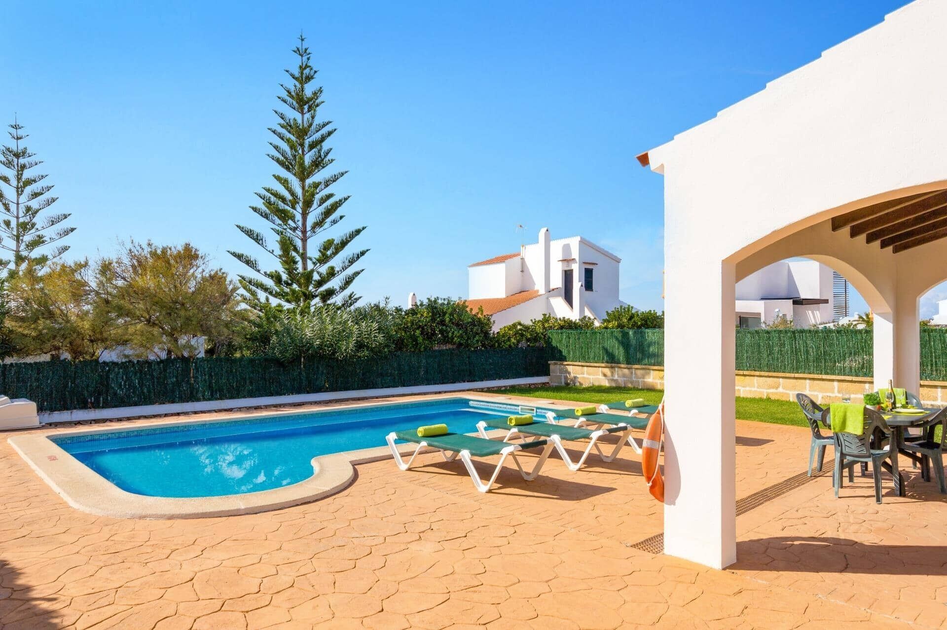 A swimming pool with lounge chairs, a covered patio area, and a white building in the background.
