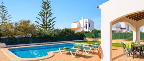 A swimming pool with lounge chairs, a covered patio area, and a white building in the background.
