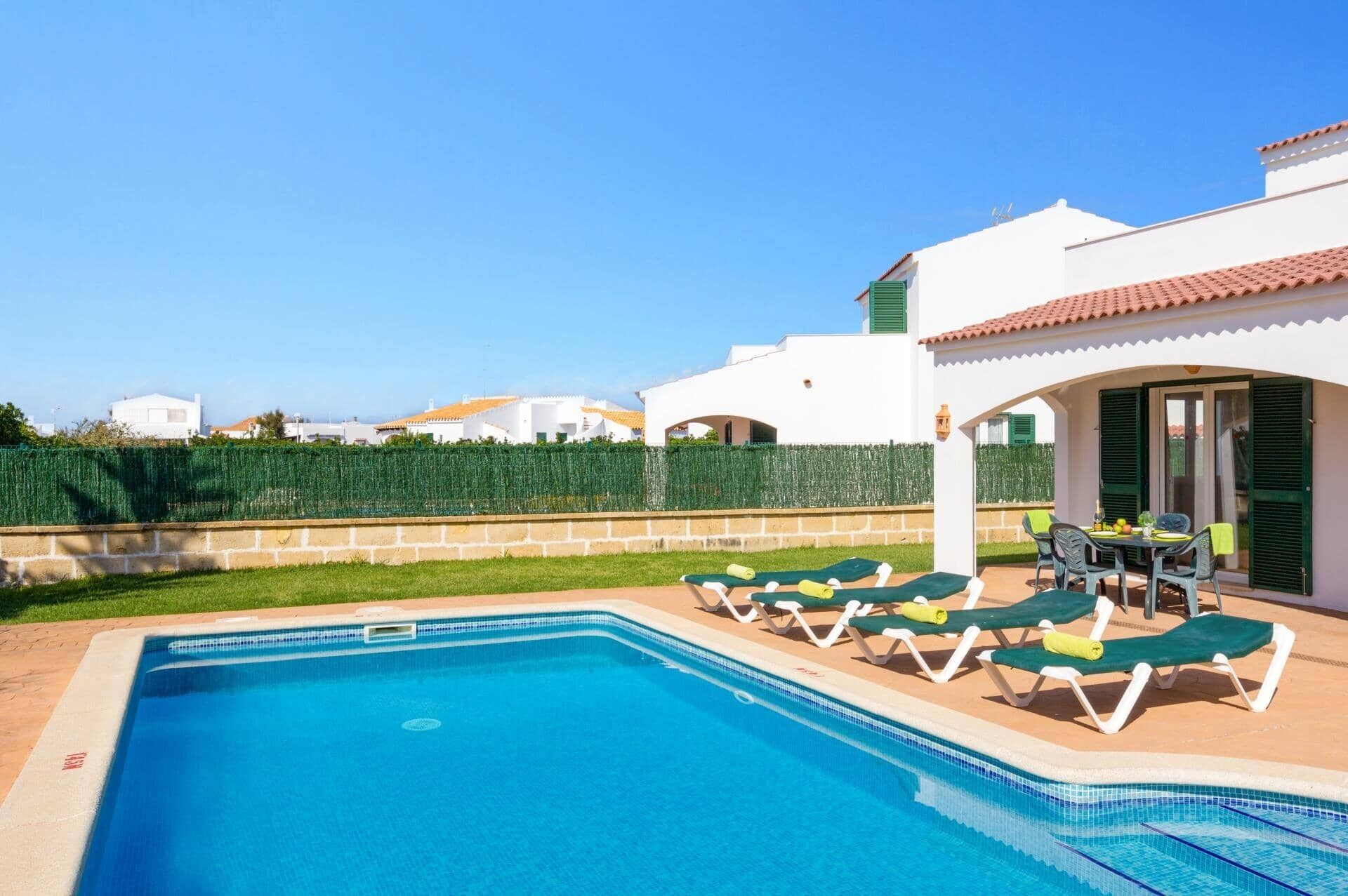 Una piscina con sillas de descanso, una mesa de patio y vistas a edificios blancos al fondo.