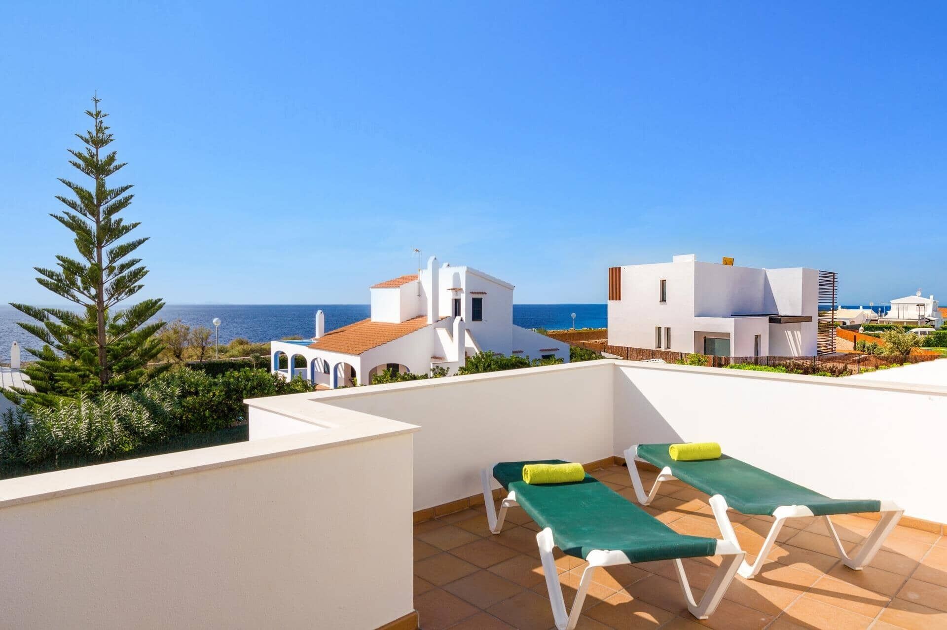 Eine Dachterrasse mit zwei grünen Liegestühlen, einem weißen Gebäude und Blick auf das Meer.