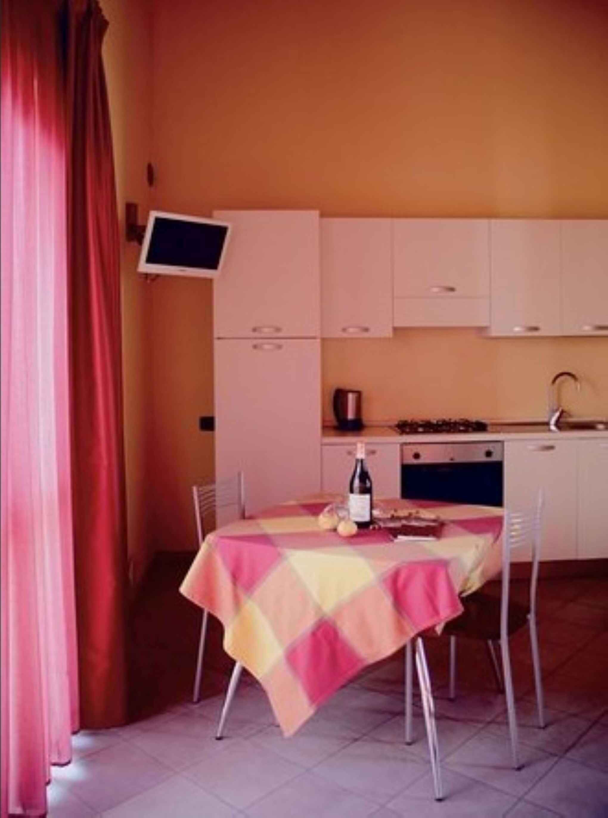 A compact kitchen with white cabinets, a dining table with a colorful tablecloth, and a TV mounted on the wall.