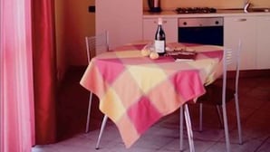 Une cuisine de taille réduite avec des meubles blancs, une table à manger recouverte d’une nappe colorée et un téléviseur fixé au mur.