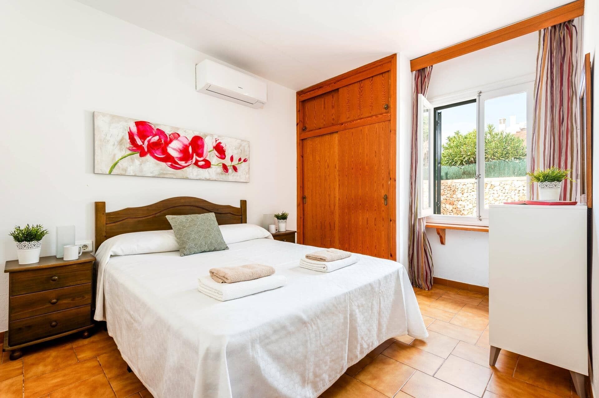 A bedroom with a wooden bed, a large window with curtains, a wooden wardrobe, and a painting of red flowers on the wall.