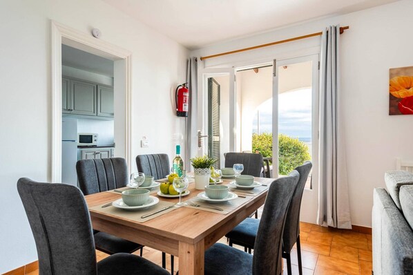 Un comedor con una mesa de madera dispuesta para cuatro personas, rodeada de sillas. Se ve una cocina a través de una puerta abierta, y un extintor está fijado a la pared.