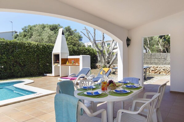 A poolside dining area with a table set for four, surrounded by white chairs, a barbecue grill, and a swimming pool.