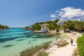 A coastal area with clear turquoise waters, a sandy beach, and buildings with balconies overlooking the sea. - Villa Salord by Villa Plus (Ciutadella de Menorca)