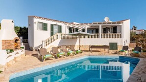 A poolside area with a pool, lounge chairs, and a building with a balcony. - Villa Santi I by Villa Plus (Cala en Porter)