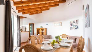 A dining area with a wooden table set for six, surrounded by wicker chairs, with a wooden cabinet and a sofa in the background. - Villa Santi I by Villa Plus (Cala en Porter)