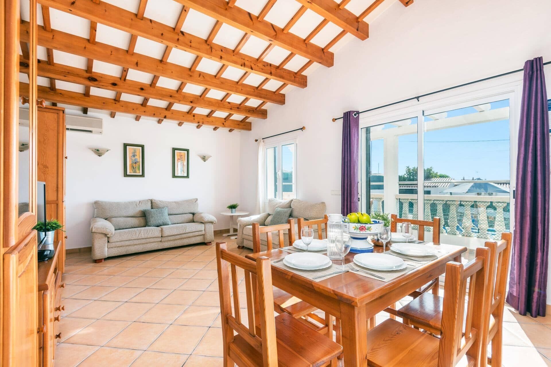 A dining area with a wooden table and chairs, a sofa, and a balcony with a view.