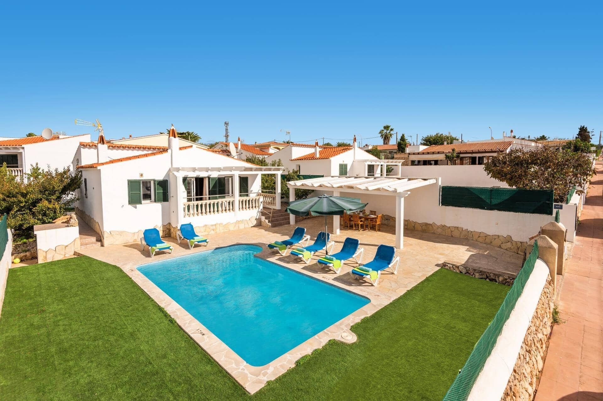 Zona de piscina con una piscina azul cristalina, rodeada por un edificio blanco con tejado de baldosas y un área de estar cubierta con mesa y sillas.