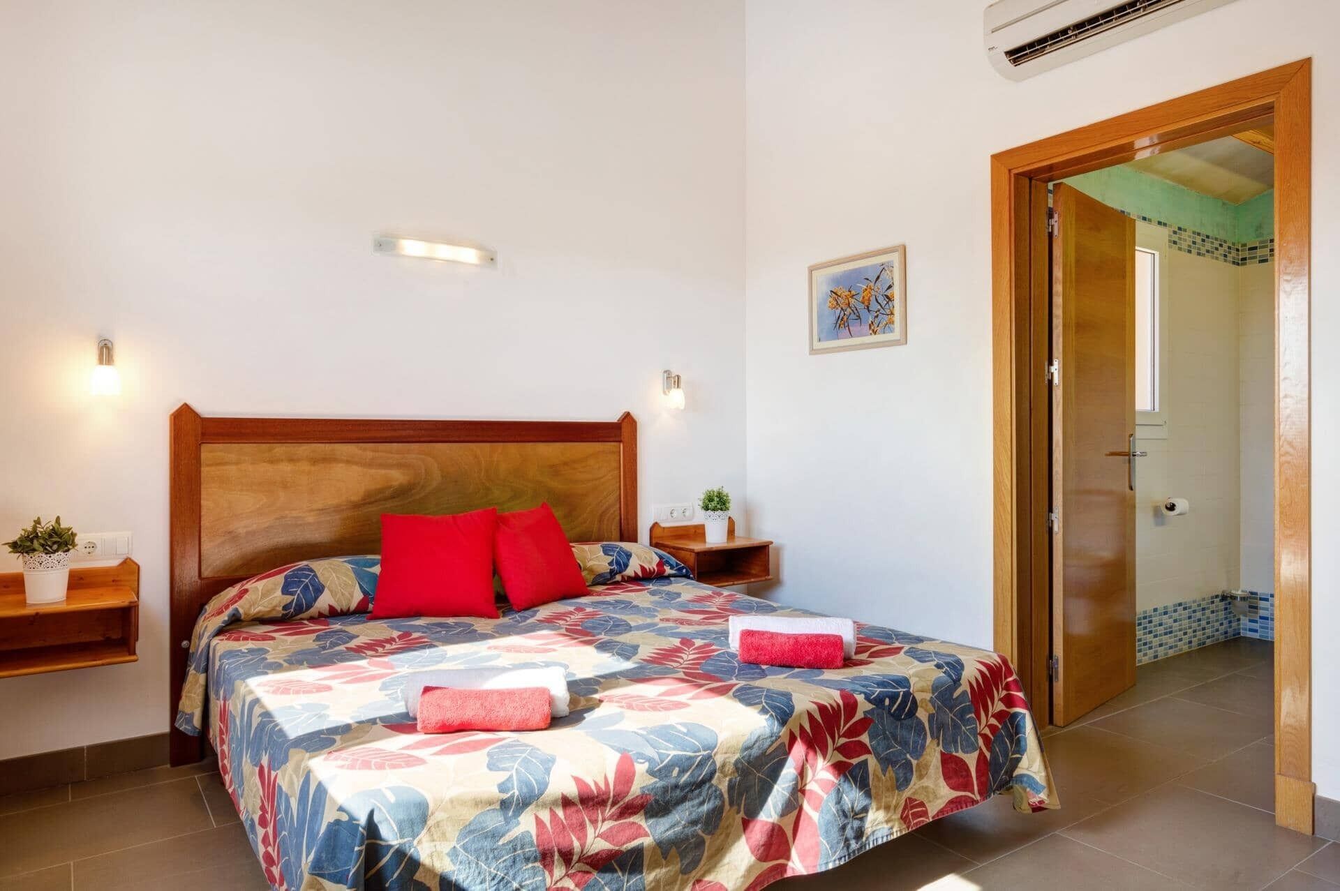 Un dormitorio con cabecera de madera, colcha estampada con motivos florales, dos almohadas rojas, una mesita de noche con una planta y una puerta que da a un baño.